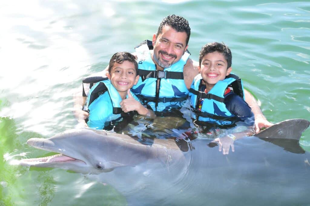 Padre con sus 2 hijos acariciando un delfín y saludando a la cámara en Cancún - Cliente de AquíViajeros